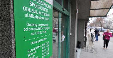 Fotografia przedstawiająca wejście do Zakładu Ubezpieczeń Społecznych przy ulicy Mickiewicza w Toruniu. Zielony szyld i drzwi wejściowe na pierwszym planie. W tle widoczny chodnik i przechodnie