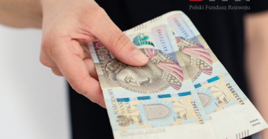 Zdjęcie przedstawia osobę trzymającą dwa banknoty 500 zł  i logotyp PFR