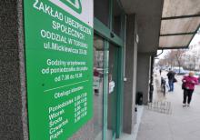 Fotografia przedstawiająca wejście do Zakładu Ubezpieczeń Społecznych przy ulicy Mickiewicza w Toruniu. Zielony szyld i drzwi wejściowe na pierwszym planie. W tle widoczny chodnik i przechodnie